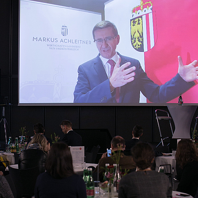 Teilnehmer des OÖ Zukunftforums 2022 sehen auf einer Leinwand die Begrüßung von Wirtschafts-Landesrat Markus Achleitner aus dem Homeoffice ©Cityfoto/Pelzl